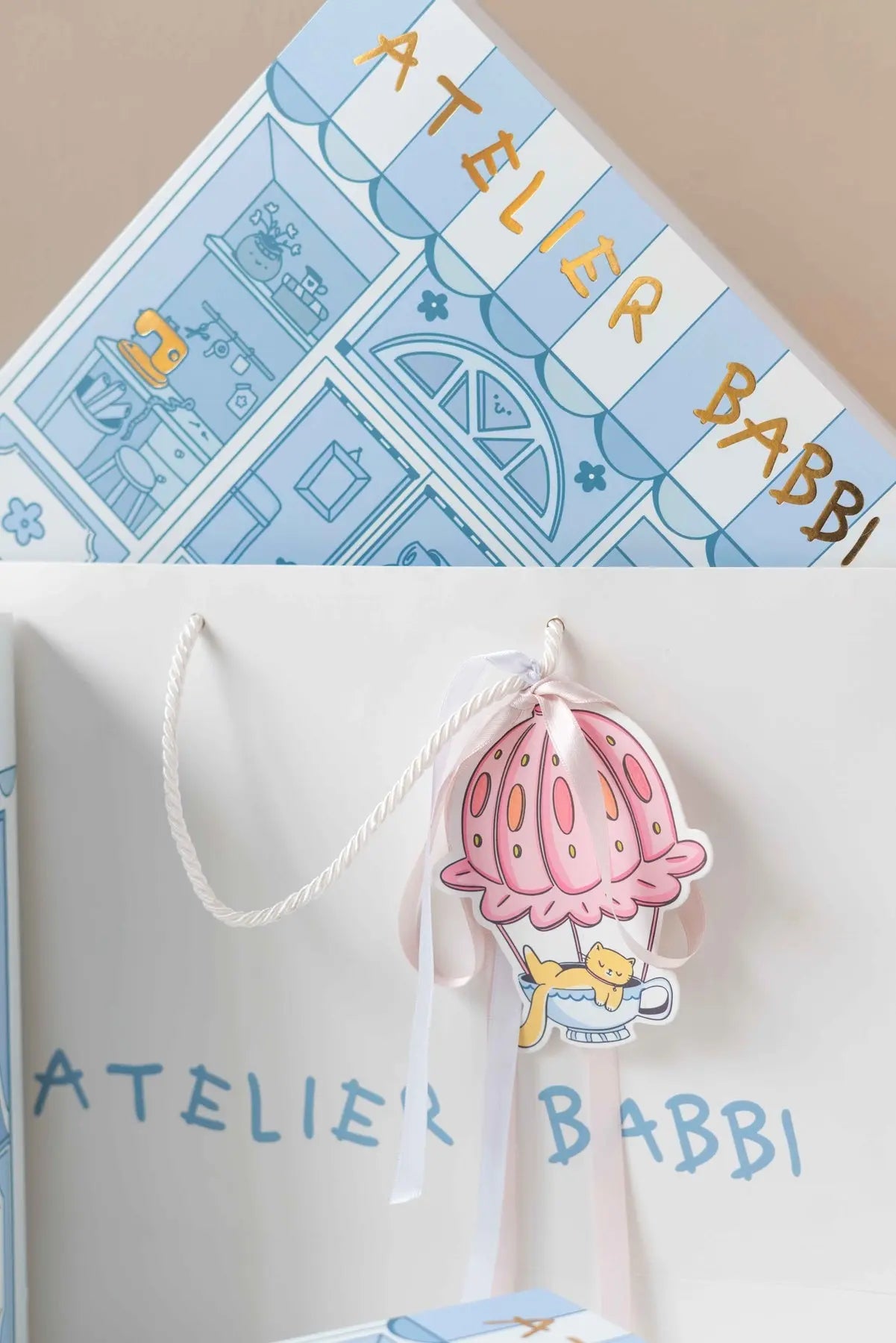 Close-up of Atelier Babbi's signature gift packaging featuring a blue illustrated boutique box and a white bag decorated with a pink hot air balloon-shaped tag and satin ribbons.