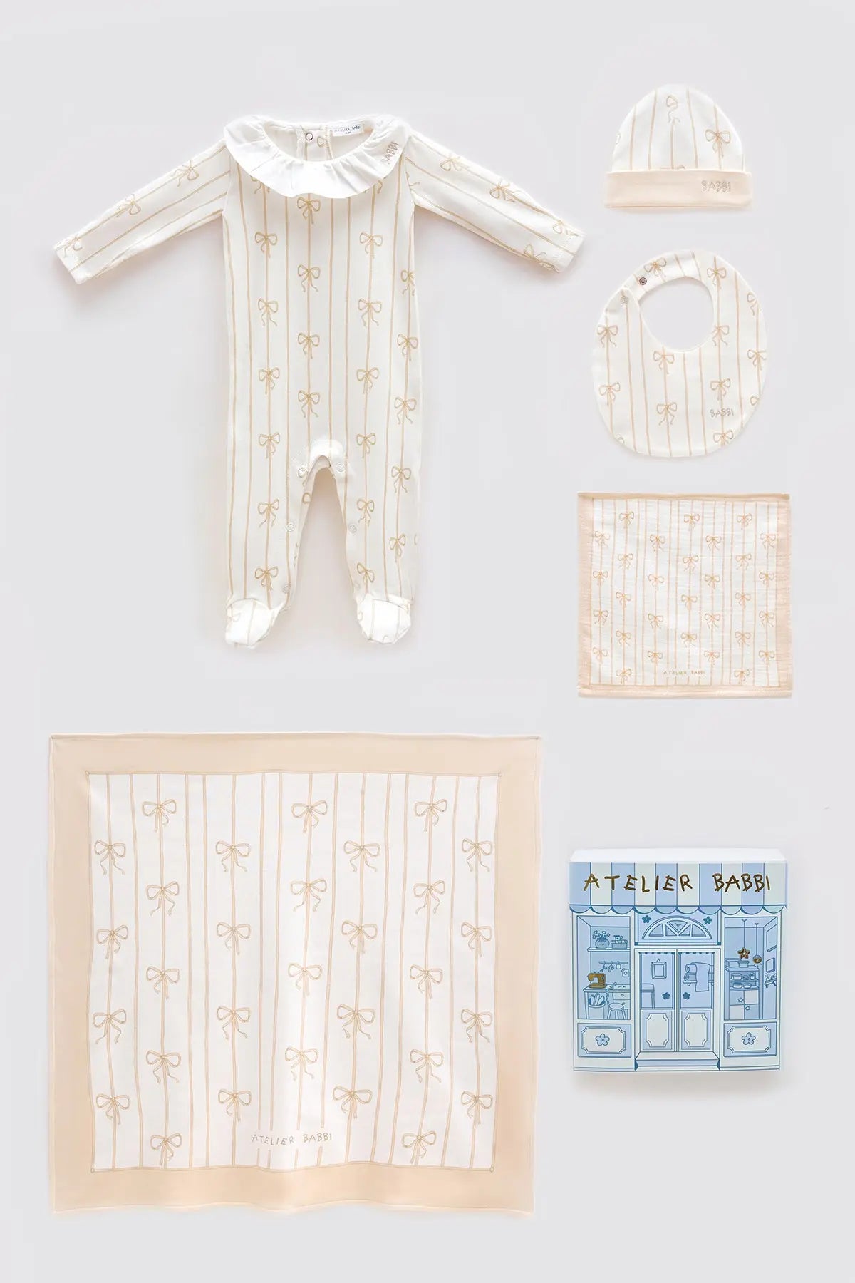 Beige Ribbon newborn gift set including a footed romper with frill collar, matching hat, bib, muslin burp cloth, and large muslin blanket, all featuring elegant beige bow patterns, styled with Atelier Babbi gift box.
