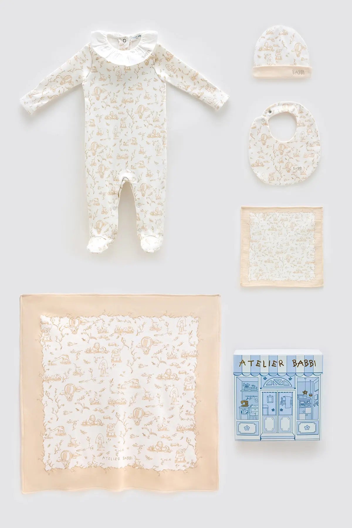 Flat lay of Atelier Babbi’s Toile de Jouy Beige 5-piece newborn set, including a footed jumpsuit with ruffled collar, matching hat, bib, muslin cloth, and a story-themed swaddle blanket, all presented with a signature gift box.