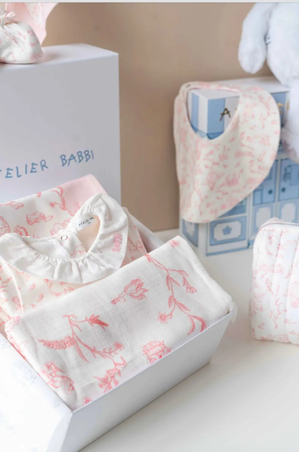 Close-up of Atelier Babbi Toile de Jouy Pink newborn gift set, featuring a folded romper with white ruffled collar, floral muslin swaddle, printed bib, and elegant packaging — all made from soft organic cotton.