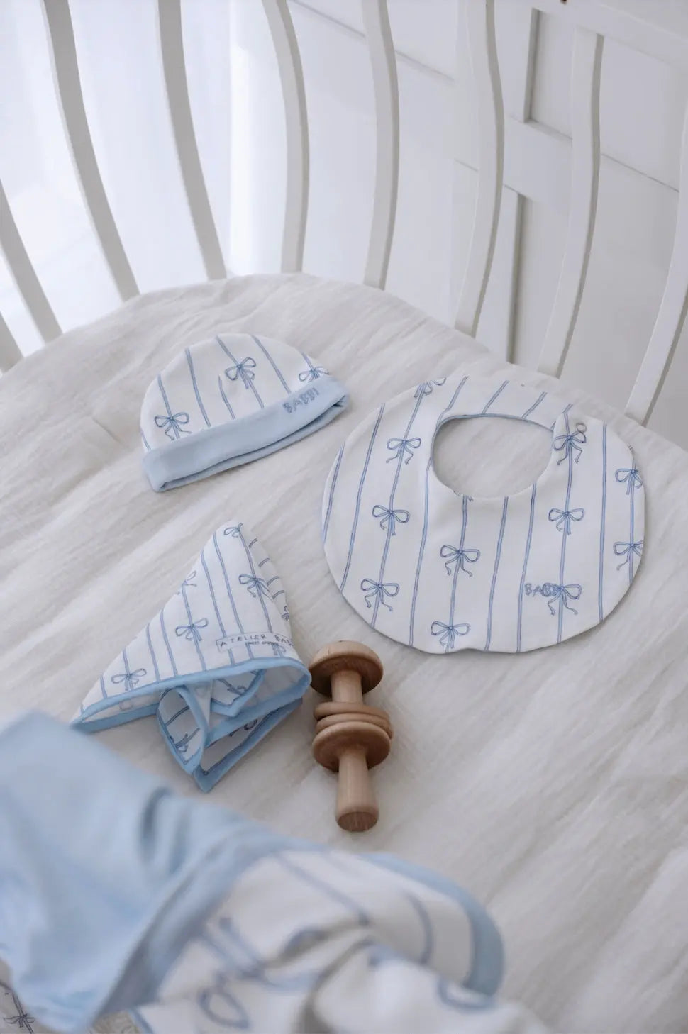 Blue Ribbon set featuring a matching baby hat, round bib, and mouth cloth neatly arranged in a white crib, accented with delicate blue bow patterns from Atelier Babbi.