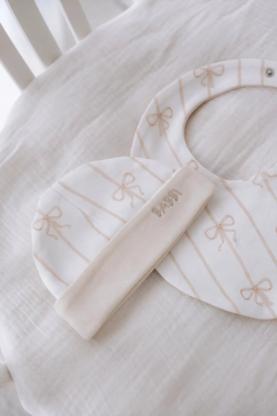 Beige ribbon patterned baby bib and hat on a soft white muslin crib sheet