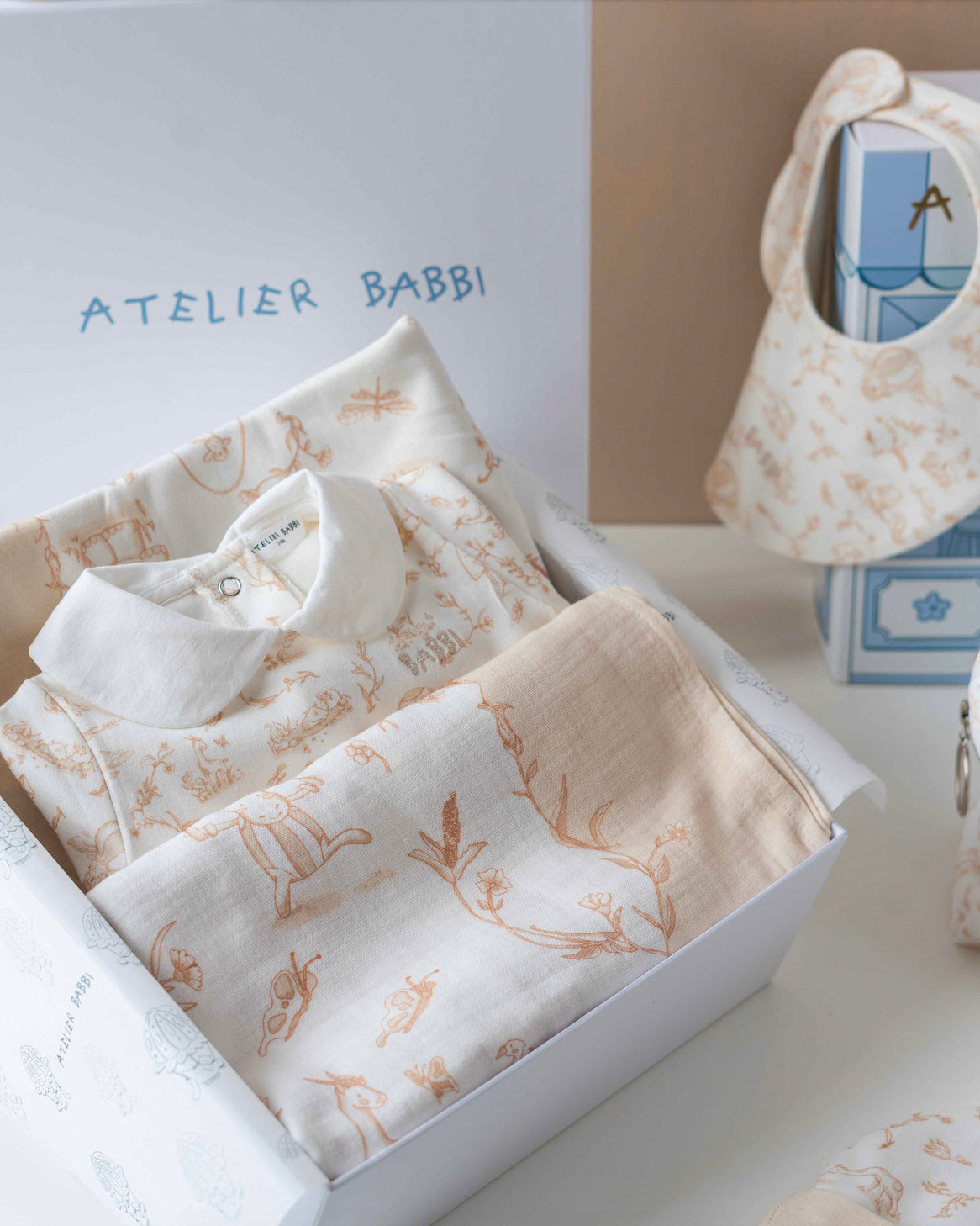 Close-up of Atelier Babbi’s Toile de Jouy Beige newborn gift set inside its premium gift box, featuring a soft cotton jumpsuit with a white collar, a matching muslin swaddle, and baby bib with delicate toile illustrations.