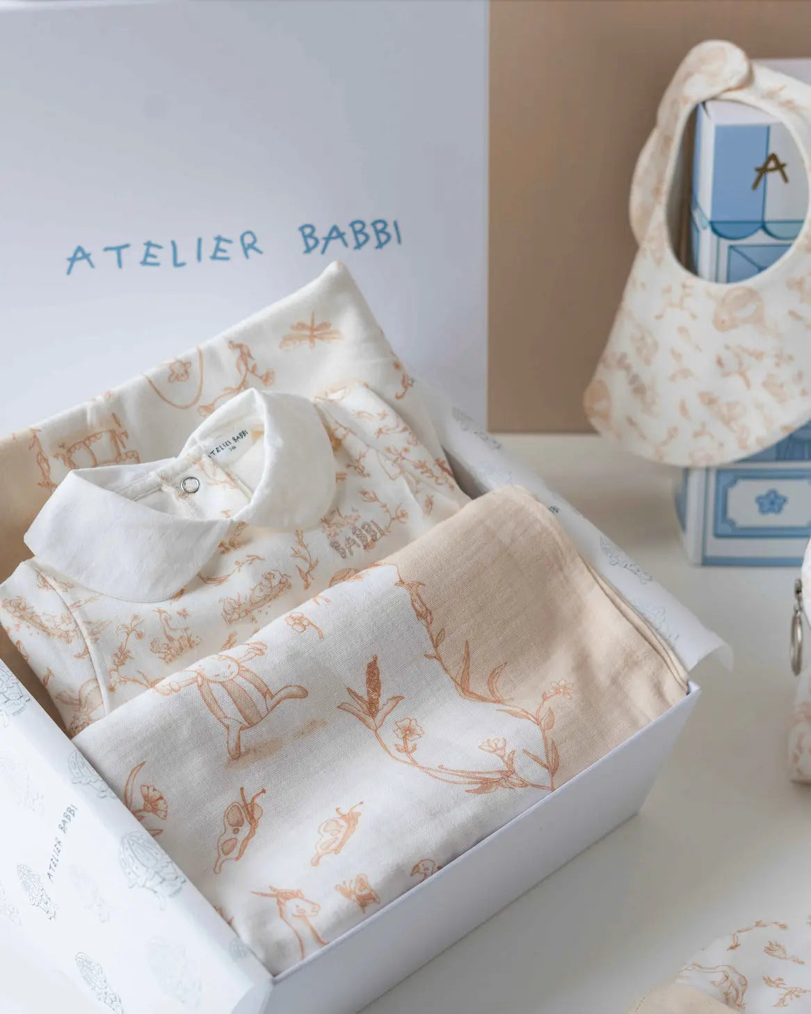Close-up of Atelier Babbi’s Toile de Jouy Beige newborn gift set inside its premium gift box, featuring a soft cotton jumpsuit with a white collar, a matching muslin swaddle, and baby bib with delicate toile illustrations.