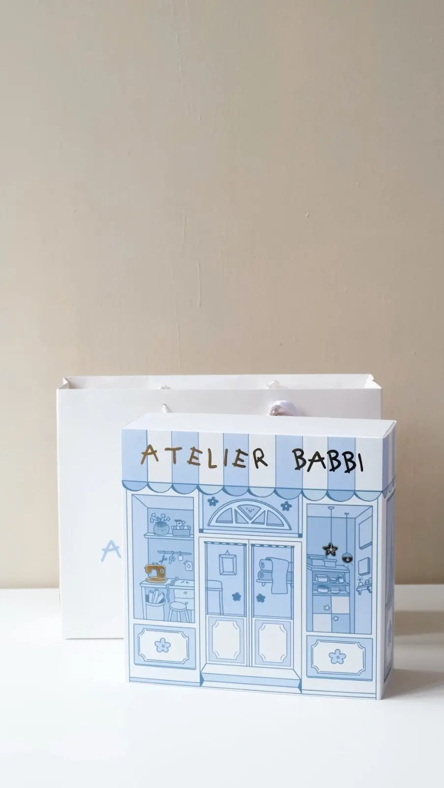 A decorative gift box with a blue illustration of a storefront on it, labeled 'Atelier Babbi'. The box is closed and presented against a beige background.