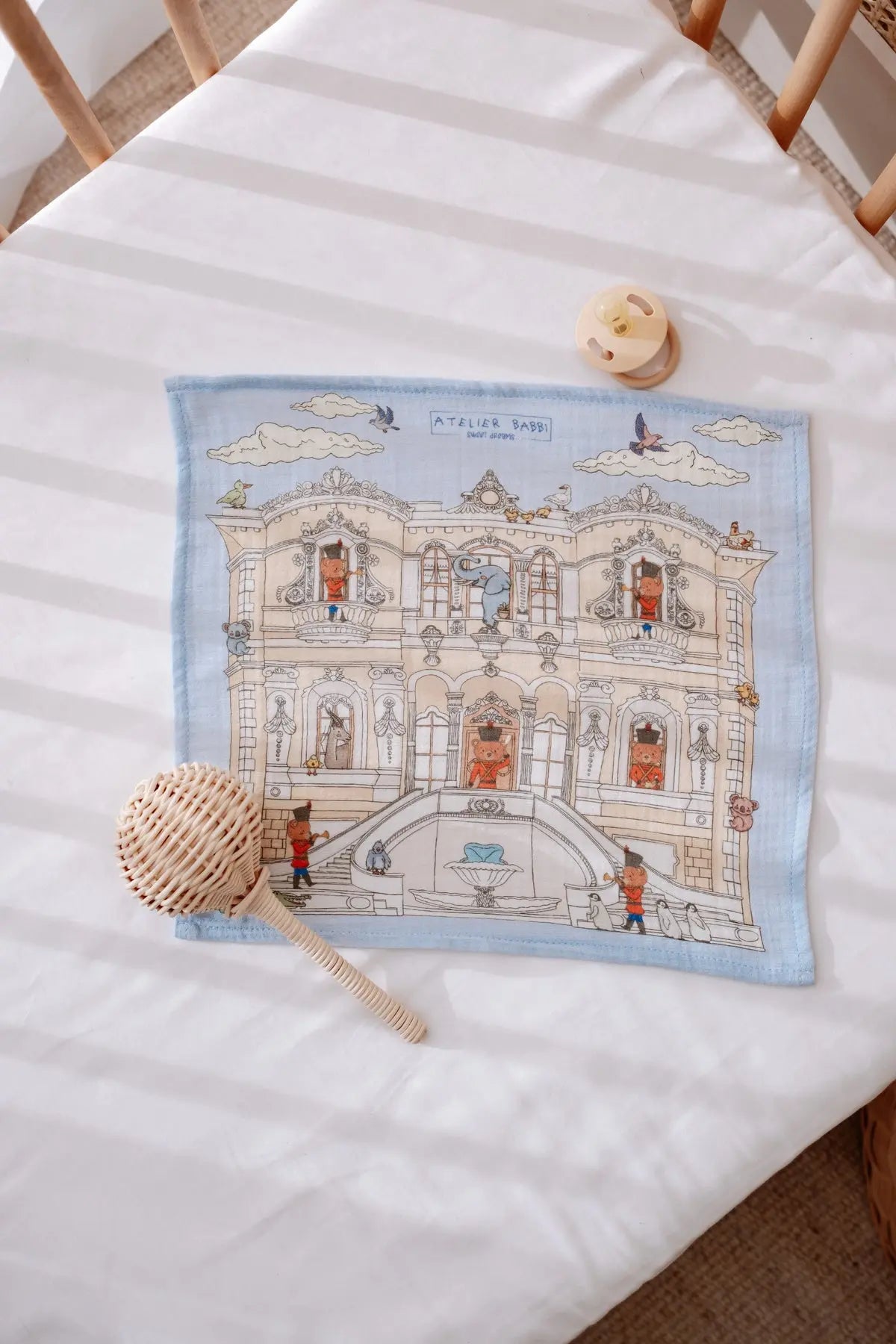 Blue-themed muslin burp cloth featuring a hand-illustrated palace with toy soldier details, laid on a white crib mattress next to a pacifier and wooden baby rattle.