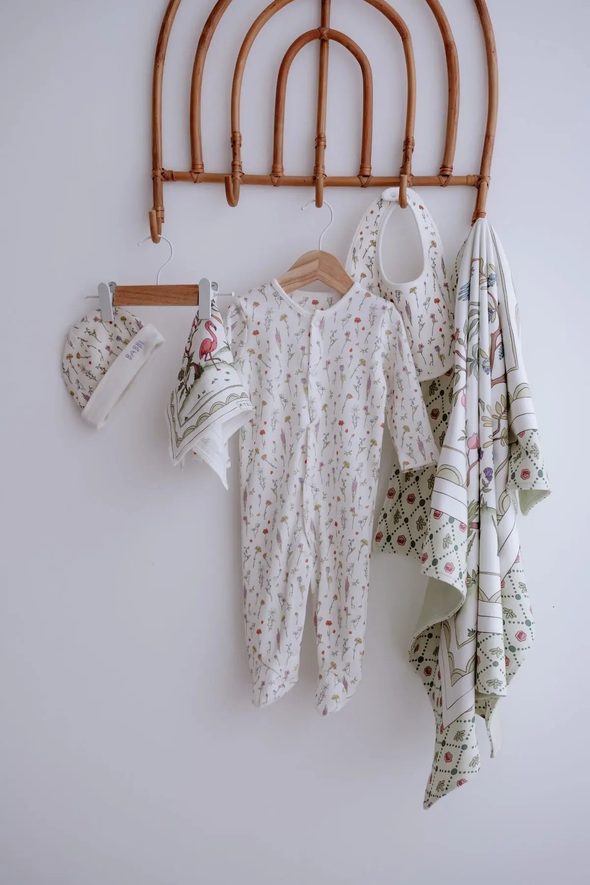 Atelier Babbi Spring Collection 5-piece baby gift set hanging on a wooden rack, featuring a floral print onesie, hat, bib, multi-use cloth, and muslin swaddle with exotic birds and botanical details — made of 100% organic cotton.
