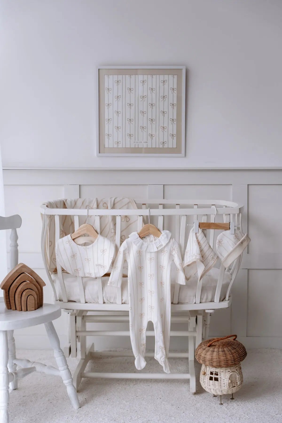 Beige Ribbon 5-piece baby gift set displayed on a white crib, including a footed romper, bib, hat, burp cloth, and muslin blanket with elegant beige bow prints, styled with a framed matching artwork.
