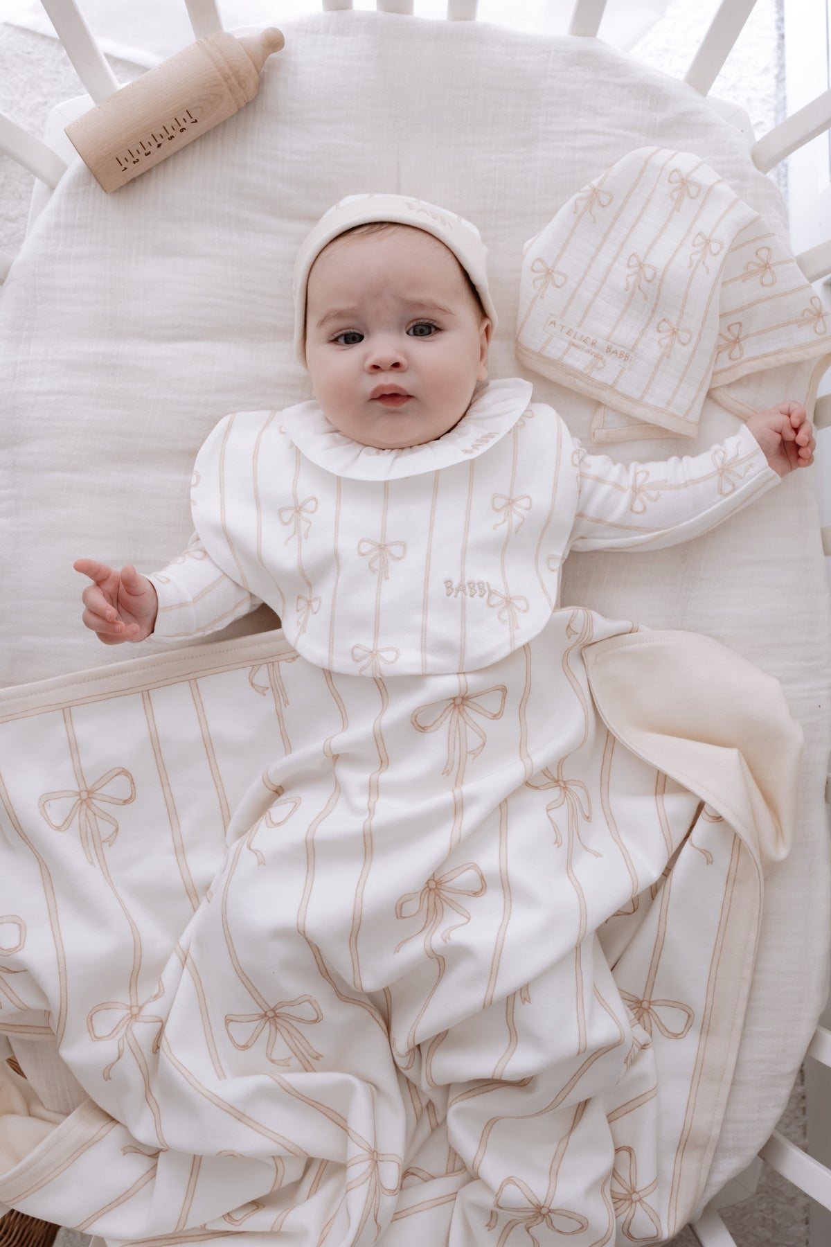 Baby lying in a crib wearing Atelier Babbi’s beige ribbon romper, matching bib and hat, wrapped in a soft cotton blanket with bow patterns — part of the Beige Ribbon collection.
