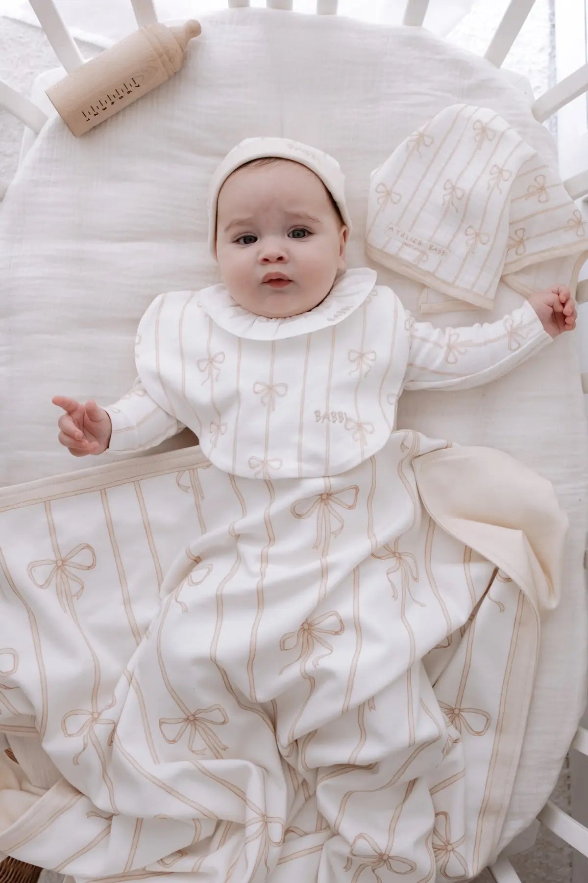Baby lying in a crib wearing Atelier Babbi’s beige ribbon romper, matching bib and hat, wrapped in a soft cotton blanket with bow patterns — part of the Beige Ribbon collection.