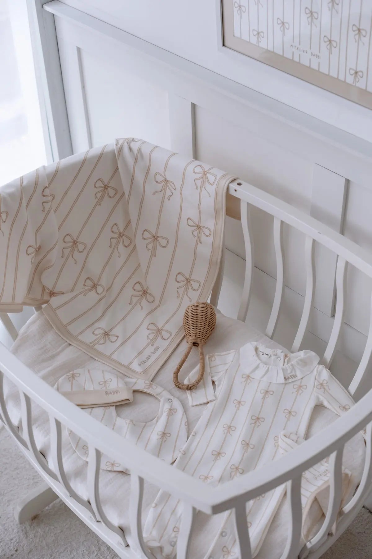 Beige ribbon patterned baby blanket, romper, bib and hat set in a white wooden crib