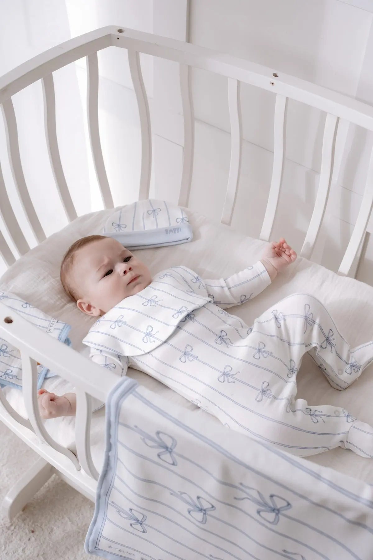 Baby lying in a white crib wearing a blue ribbon patterned onesie, matching bib and hat from the Atelier Babbi Blue Ribbon newborn set, surrounded by coordinated nursery textiles.