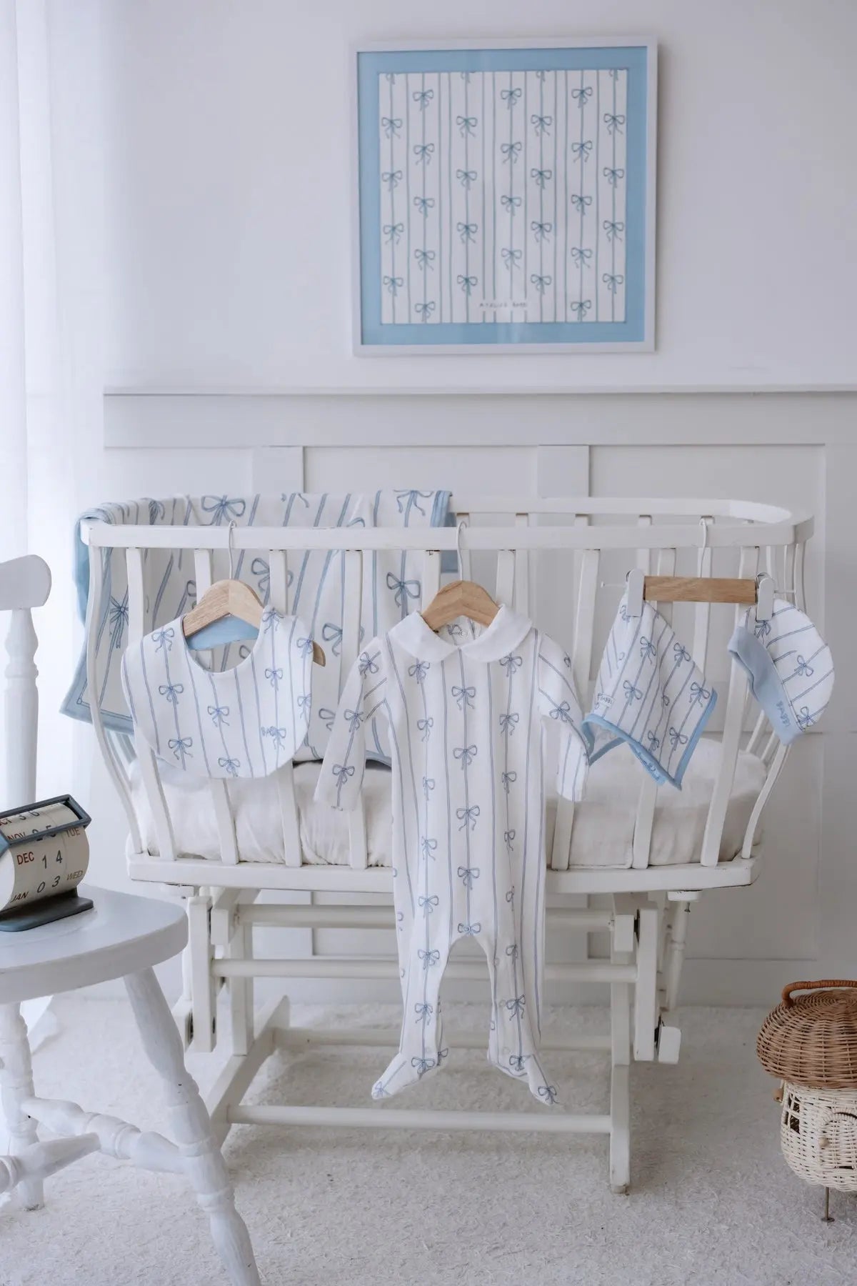 Blue Ribbon themed 5-piece newborn set displayed on a white cradle, featuring an organic cotton onesie, bib, hat, swaddle blanket, and burp cloth with elegant blue bow patterns