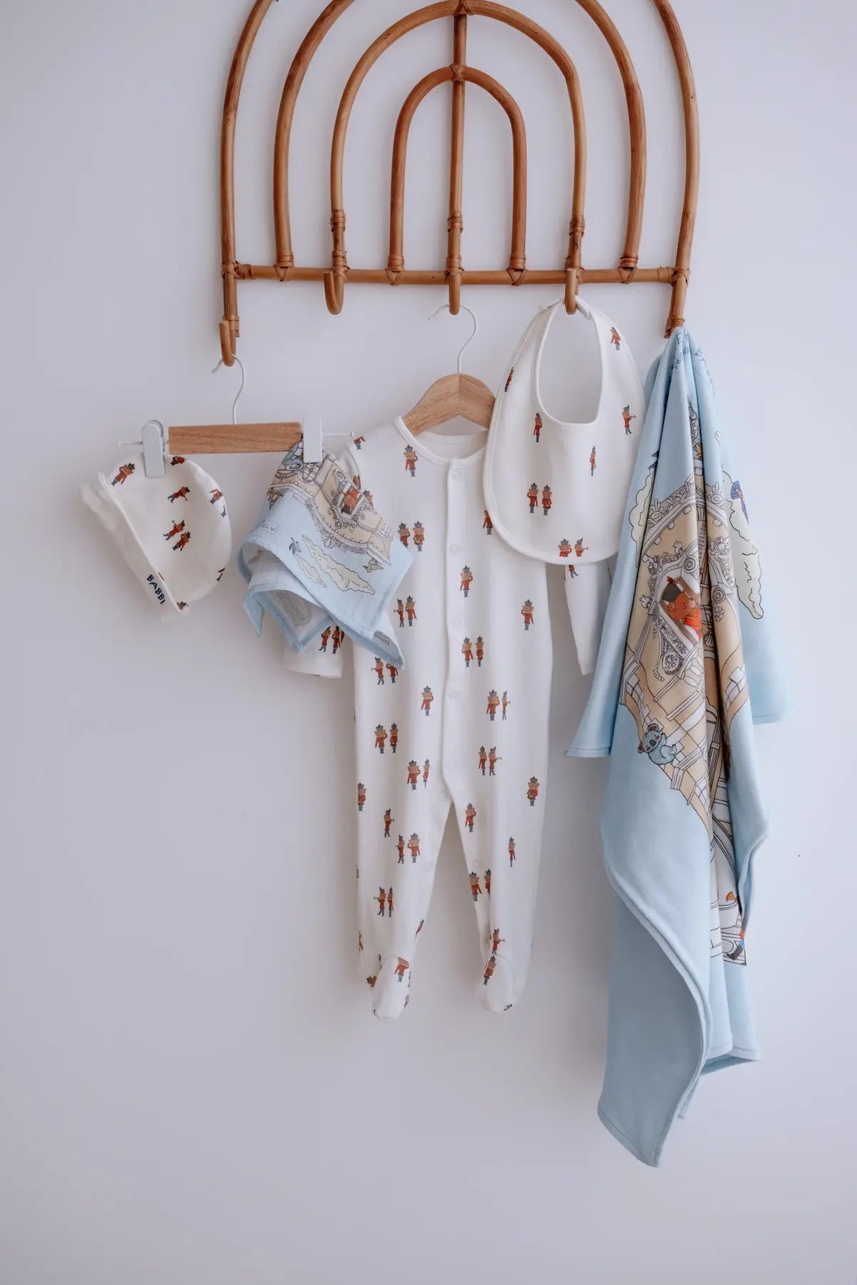 Newborn baby outfit set in Tin Soldier theme, featuring a printed romper, matching hat, bib, muslin cloth, and soft blanket with palace illustration, displayed on wooden hangers against a white wall.