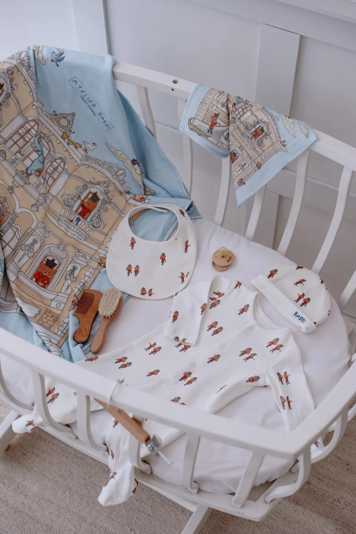 Tin Soldier newborn gift set displayed in a white crib, featuring a romper, bib, hat, and muslin cloth with a whimsical palace print in soft blue and beige tones.