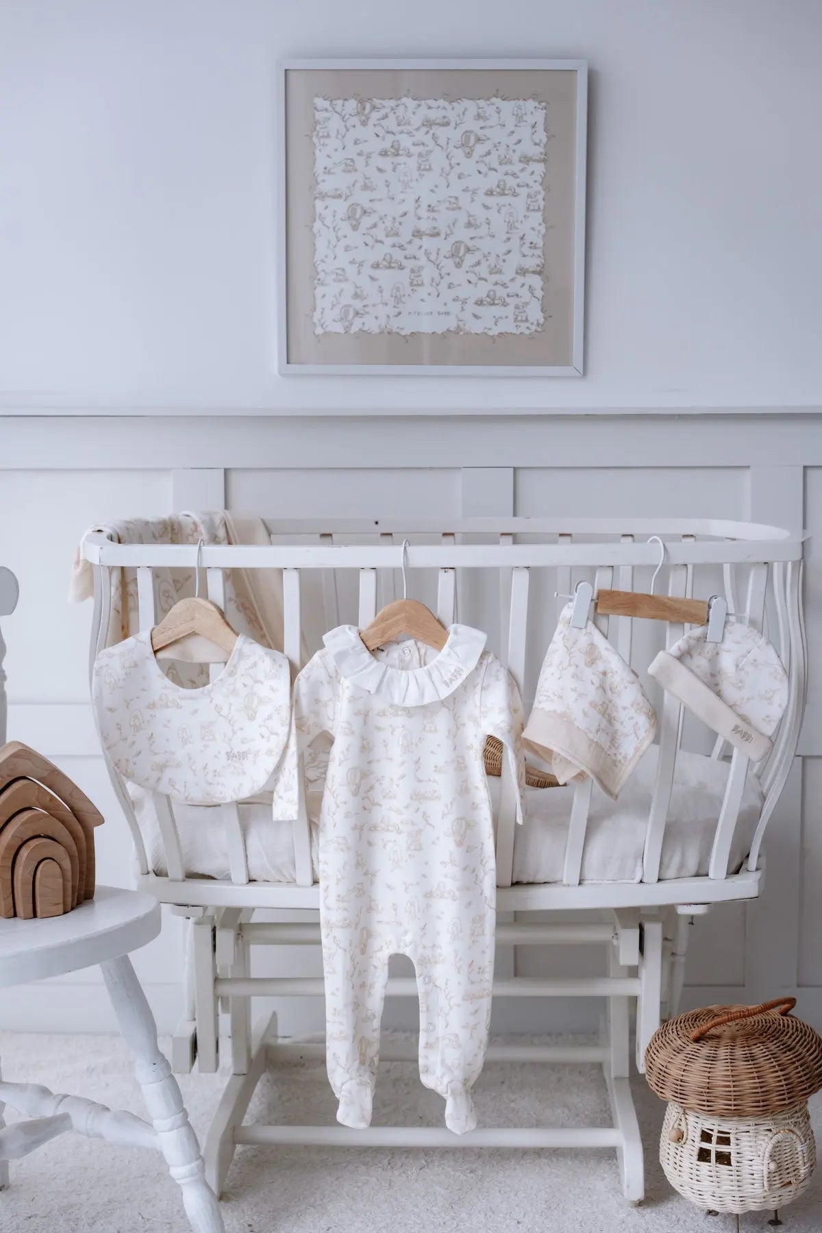 Atelier Babbi Toile de Jouy Beige 5-piece newborn gift set displayed on a white vintage crib, featuring a footed jumpsuit with white collar, matching bib, hat, muslin swaddle, and burp cloth with whimsical beige illustrations.