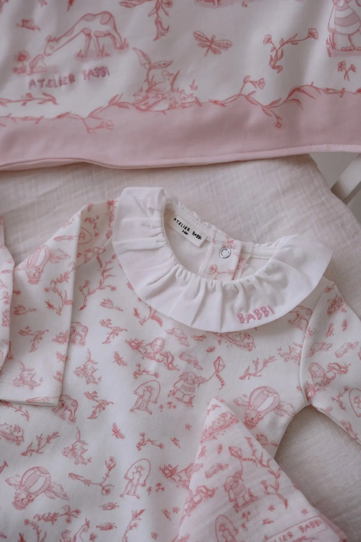 Close-up of the Toile de Jouy Pink romper with white ruffled collar, featuring whimsical illustrations and delicate embroidery, displayed with a matching muslin cloth and blanket corner.