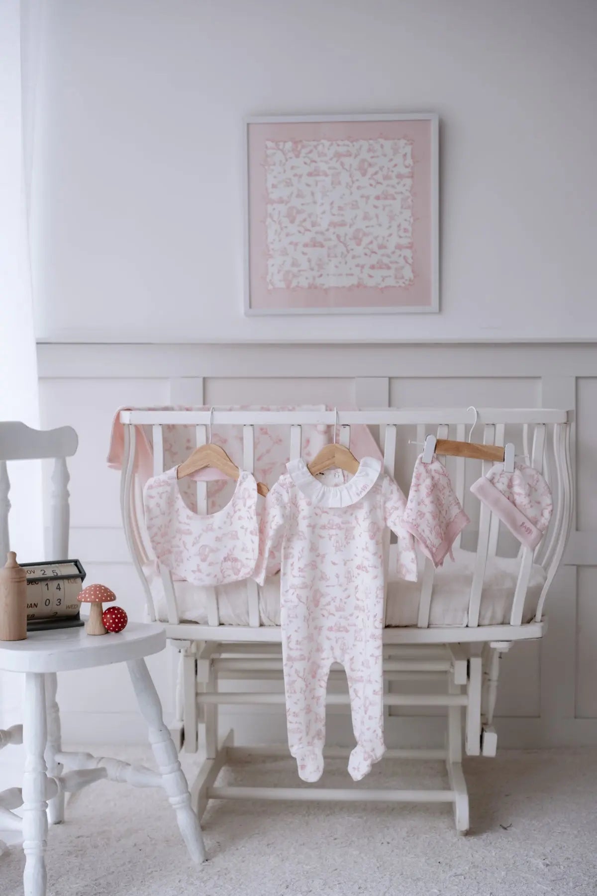 Toile de Jouy Pink 5-piece newborn set displayed on a white crib, featuring a ruffled romper, bib, hat, muslin swaddle, and framed matching textile art — a soft and elegant nursery presentation.